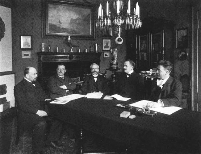 Schwarz-Weiß-Fotografie von fünf Männern, die an einem Tisch unter einem Kronleuchter sitzen und Dokumente vor sich ausgebreitet haben