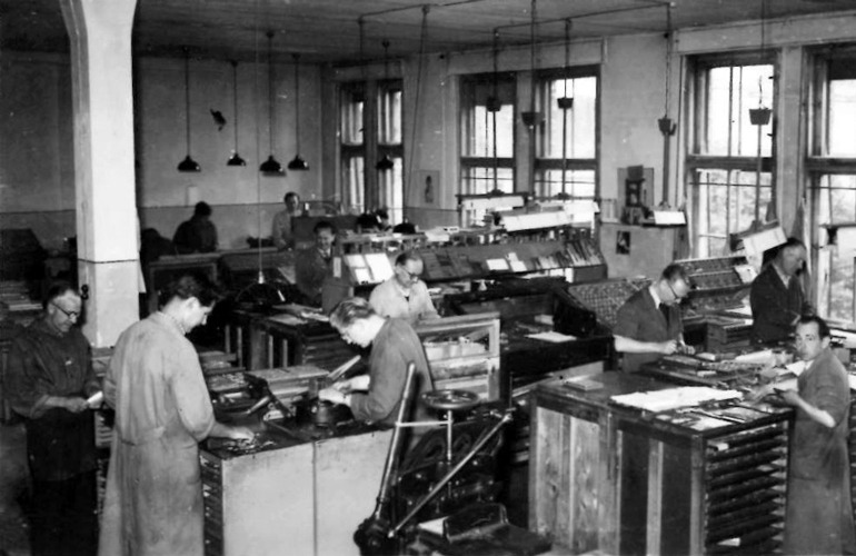 Black and white photograph of a room full of men in work coats arranging type