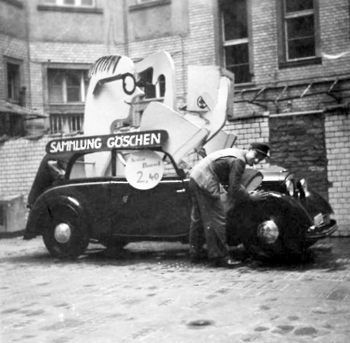 Schwarz-Weiß-Foto einer Mercedes-Benz-Limousine mit einer großen Pappfigur und der Aufschrift „Sammlung Göschen“ auf dem Dach; ein Mann mit Schiebermütze und Zigarette im Mund beugt sich über das Auto